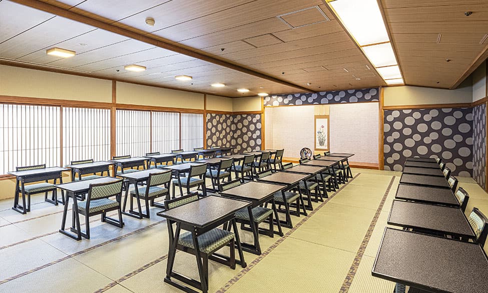 畳敷きの和室で、イスとテーブルが並べられたレイアウトの宴会場。窓から庭園が見える開放感のある空間。